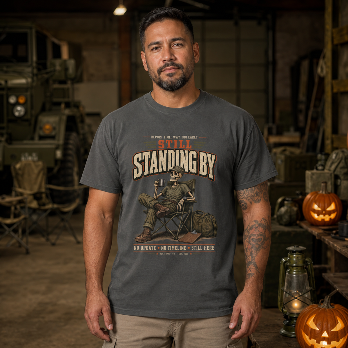 A man wearing a dark gray graphic t-shirt with a vintage-style illustration and the text 'STILL STANDING BY' in a rustic, military-themed setting with pumpkins and an olive military vehicle in the background.