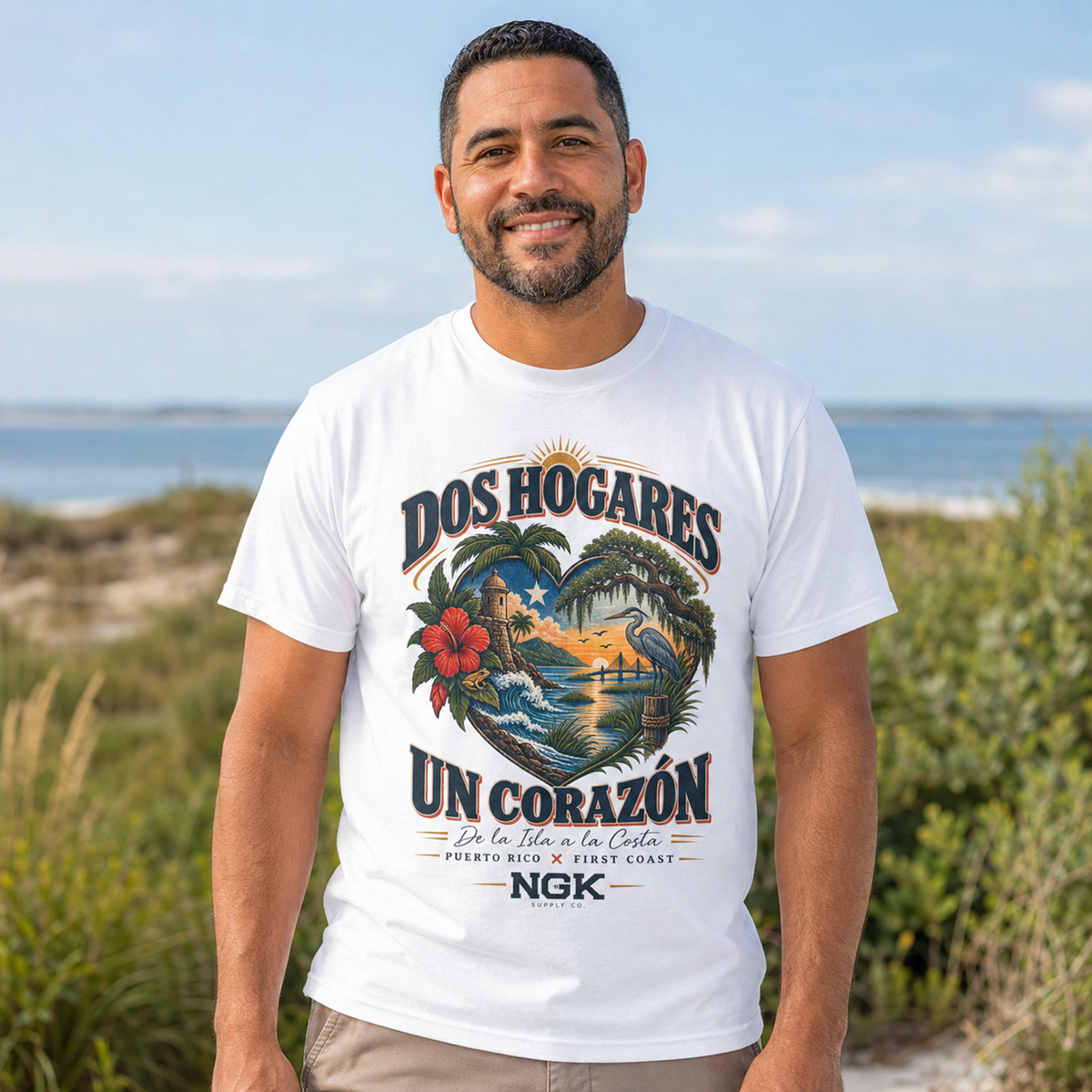 A man wearing a white graphic t-shirt with a colorful tropical design and Spanish text, standing outdoors on a beach dune with the ocean in the background.