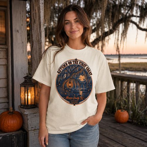 A woman stands on a wooden porch at sunset wearing an off-white graphic T-shirt with a circular blue and orange illustration and the text 'Live to Tell the Fish Catch' and 'Mistral County FL.' Pumpkins and lanterns are visible on the porch, with water and trees in the background.
