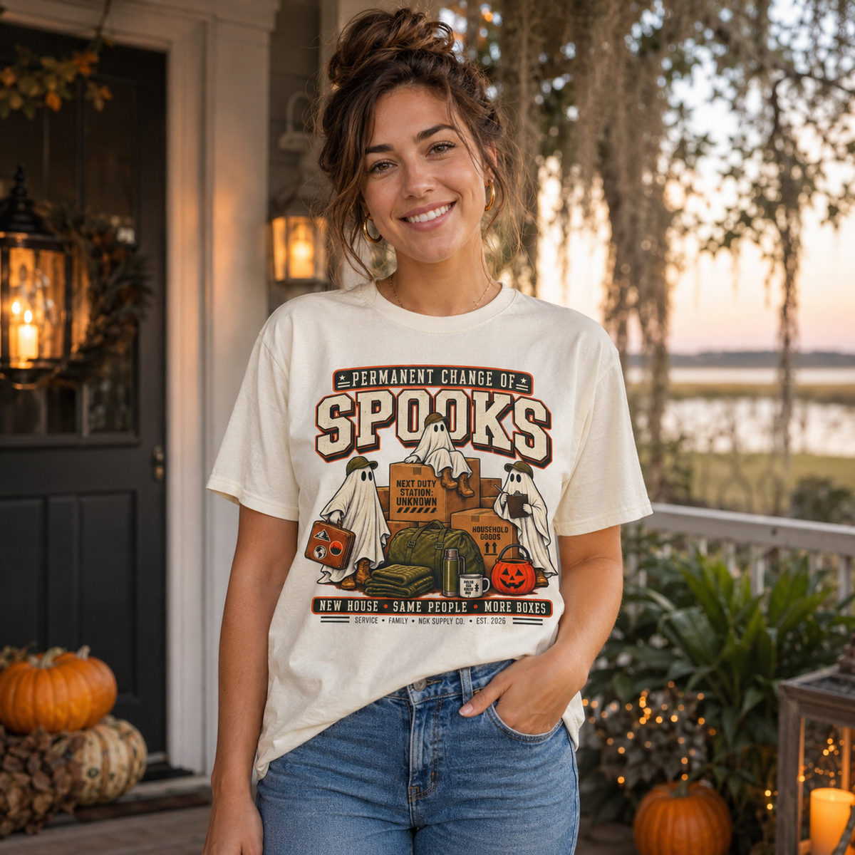 A woman stands on a decorated porch wearing a cream graphic T-shirt with a Halloween-themed print reading 'Permanent Change of Spooks' and blue jeans. Pumpkins, lanterns, plants, and a softly blurred outdoor background are visible.