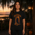 A woman stands on a wooden deck at sunset wearing a black graphic T-shirt with orange and yellow artwork and the text 'EL CUCO Ya Vienes Nick.' The background shows water, trees, lanterns, and pumpkins.