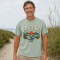 A smiling man wearing a sage green graphic t-shirt with a 'Low Tide Local' sunset and river scene print, standing on a sandy beach path with dune grass in the background.