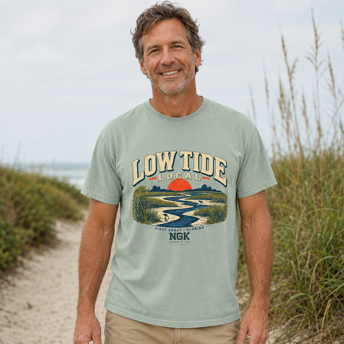 A smiling man wearing a sage green graphic t-shirt with a 'Low Tide Local' sunset and river scene print, standing on a sandy beach path with dune grass in the background.