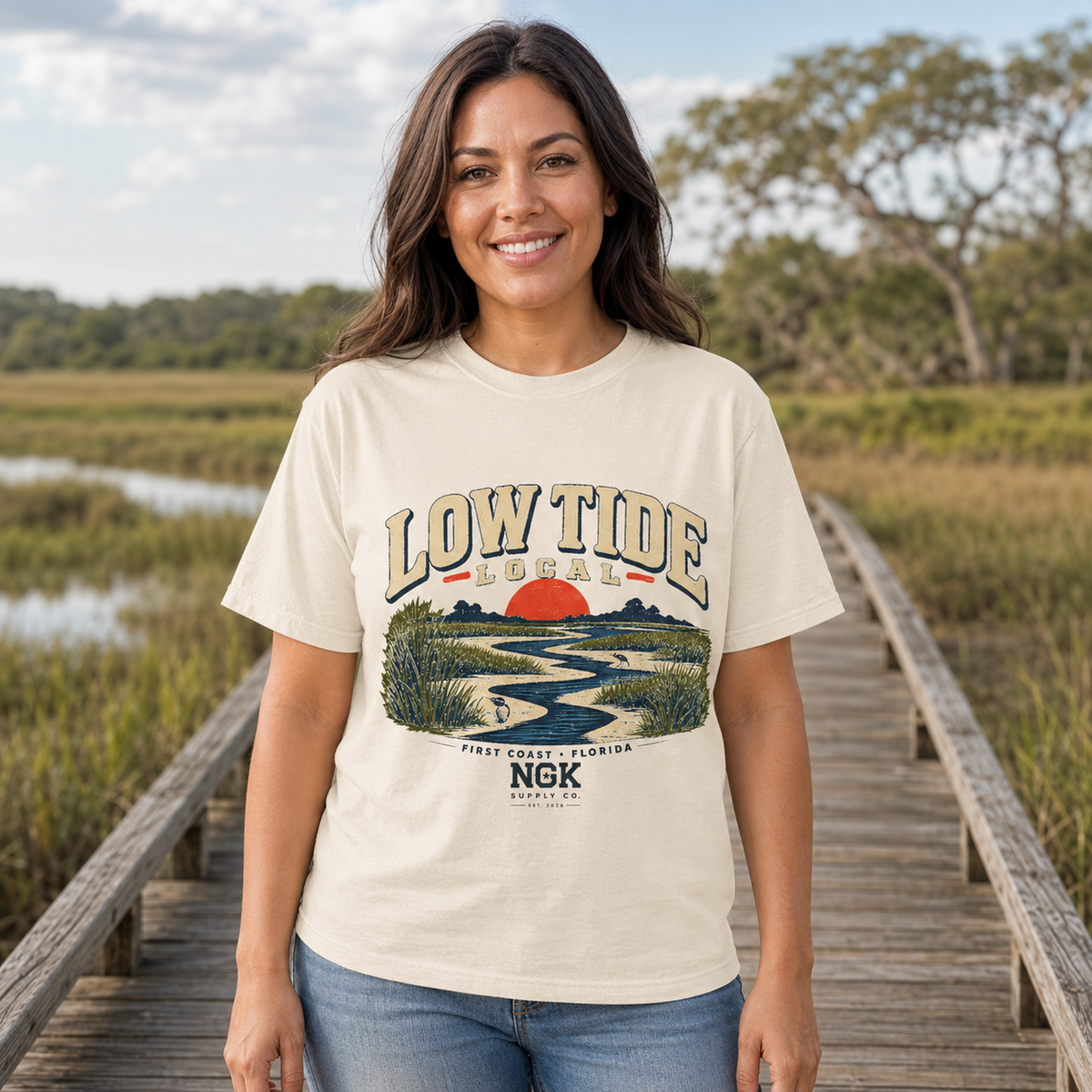 A woman stands on a wooden boardwalk in a marshy outdoor setting wearing a cream-colored graphic t-shirt that reads 'LOW TIDE LOCAL' with a sunset landscape illustration and 'NGK' text.