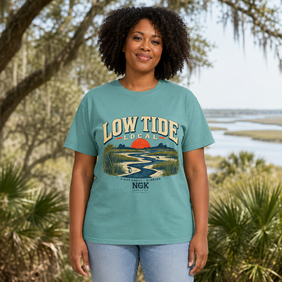 A woman wearing a teal graphic t-shirt with a 'Low Tide Local' Florida landscape print, standing outdoors in front of a blurred marsh and trees background.