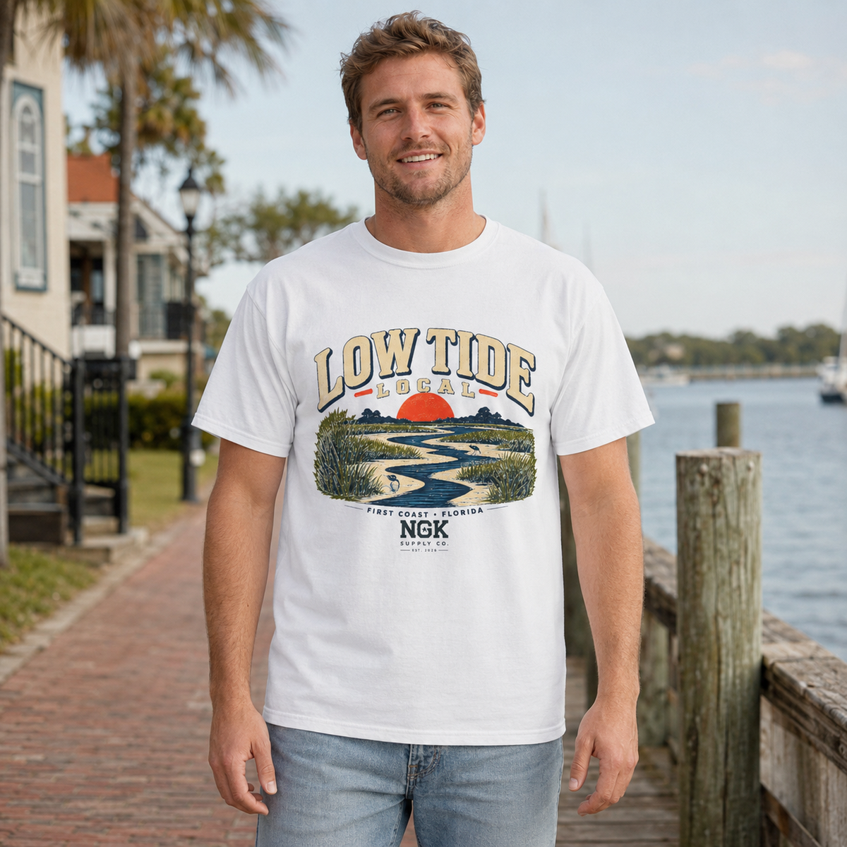 A man wearing a white graphic t-shirt with a 'Low Tide Coastal' landscape print, standing on a wooden dock next to the water.