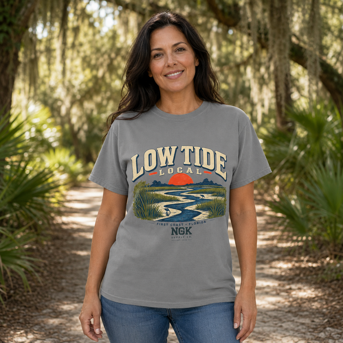 A woman stands on a sunlit outdoor path wearing a heather gray graphic T-shirt with a retro-style landscape print and the words “LOW TIDE LOCAL” across the chest. The shirt also includes smaller text reading “FLORIDA WATERFRONT EST.