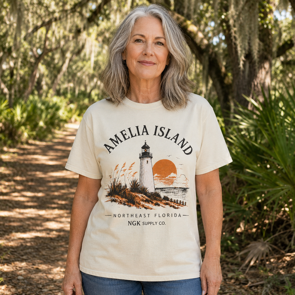 A woman with gray hair stands on a sun-dappled forest path wearing a cream-colored graphic t-shirt that reads 'AMELIA ISLAND NORTHEAST FLORIDA NGK SUPPLY CO.' with a lighthouse and sunset illustration.
