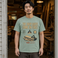 A man wearing a sage green graphic t-shirt with a vintage-style woodshop print, standing in a real workshop environment.
