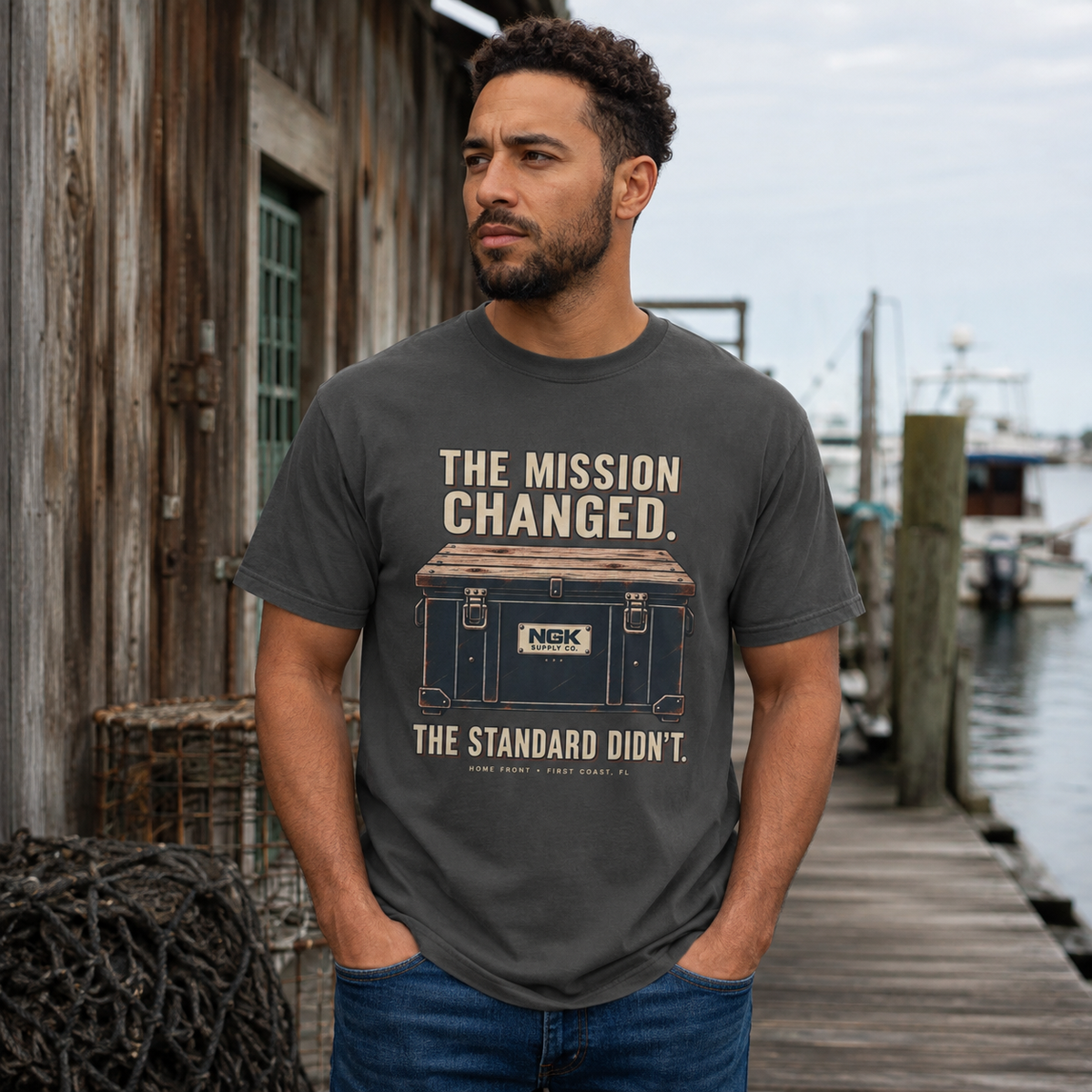 A man wearing a dark gray graphic t-shirt stands on a wooden dock next to a rustic wooden building. The t-shirt features a vintage-style illustration of a blue cooler with the text 'THE MISSION CHANGED.