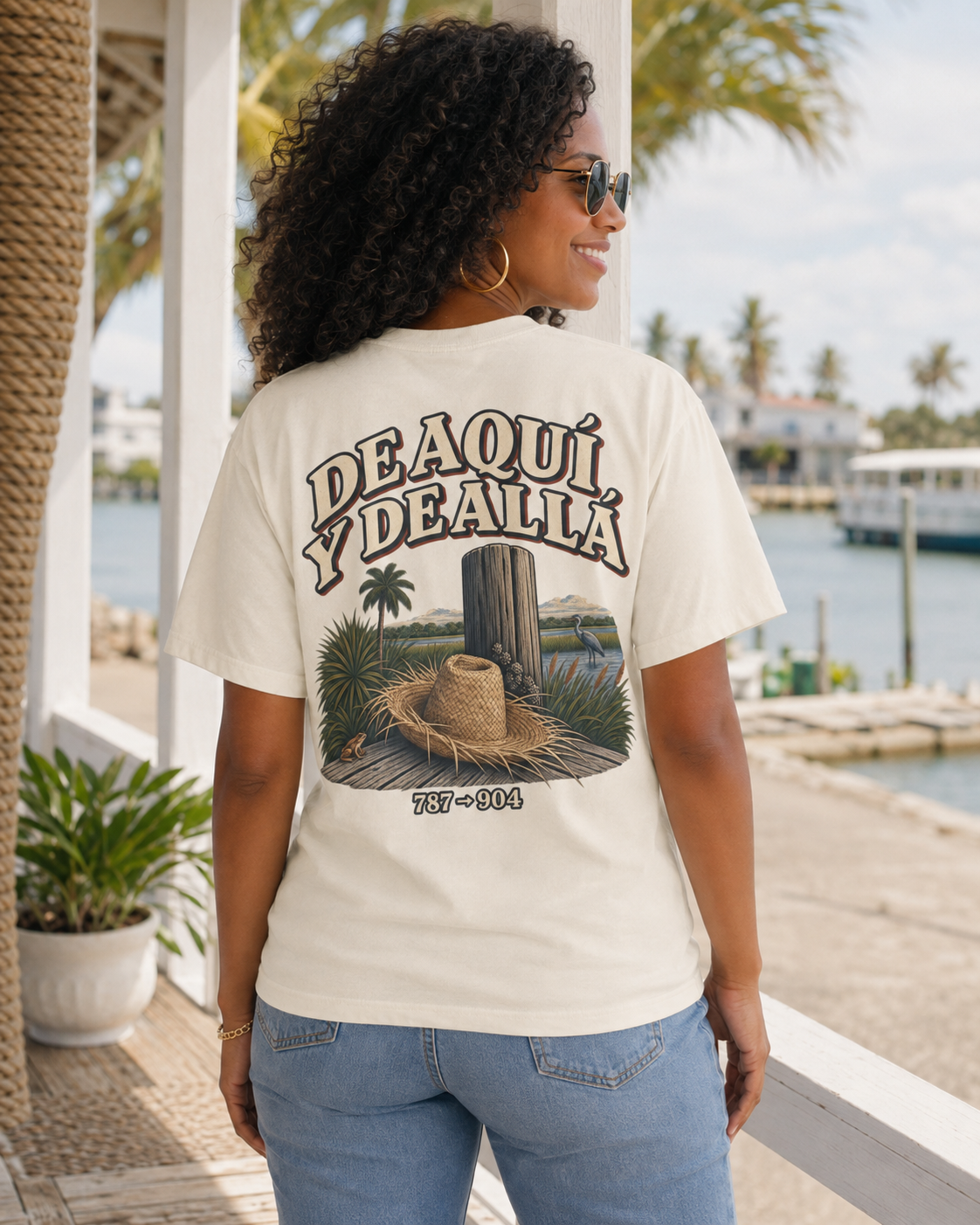 A woman wearing a cream-colored graphic t-shirt with the text 'DEAQUÍ Y DEALLÁ' and a cactus/hat illustration, shown from the back in an outdoor waterfront setting.