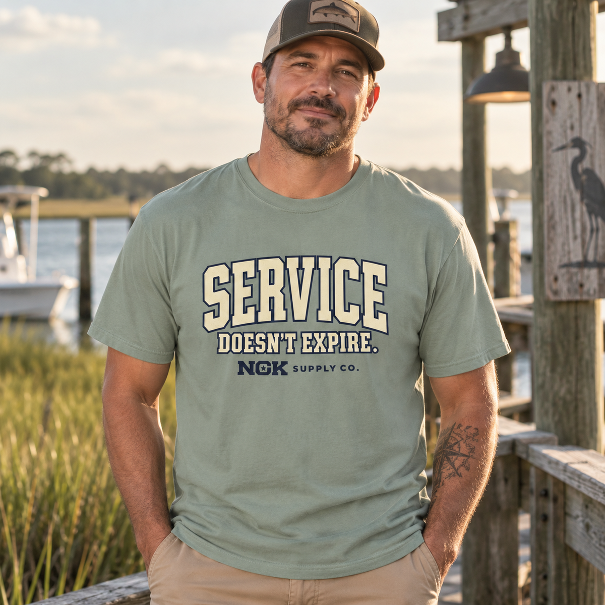 A man wearing a sage green t-shirt with 'SERVICE DOESN'T EXPIRE. NGK SUPPLY CO.