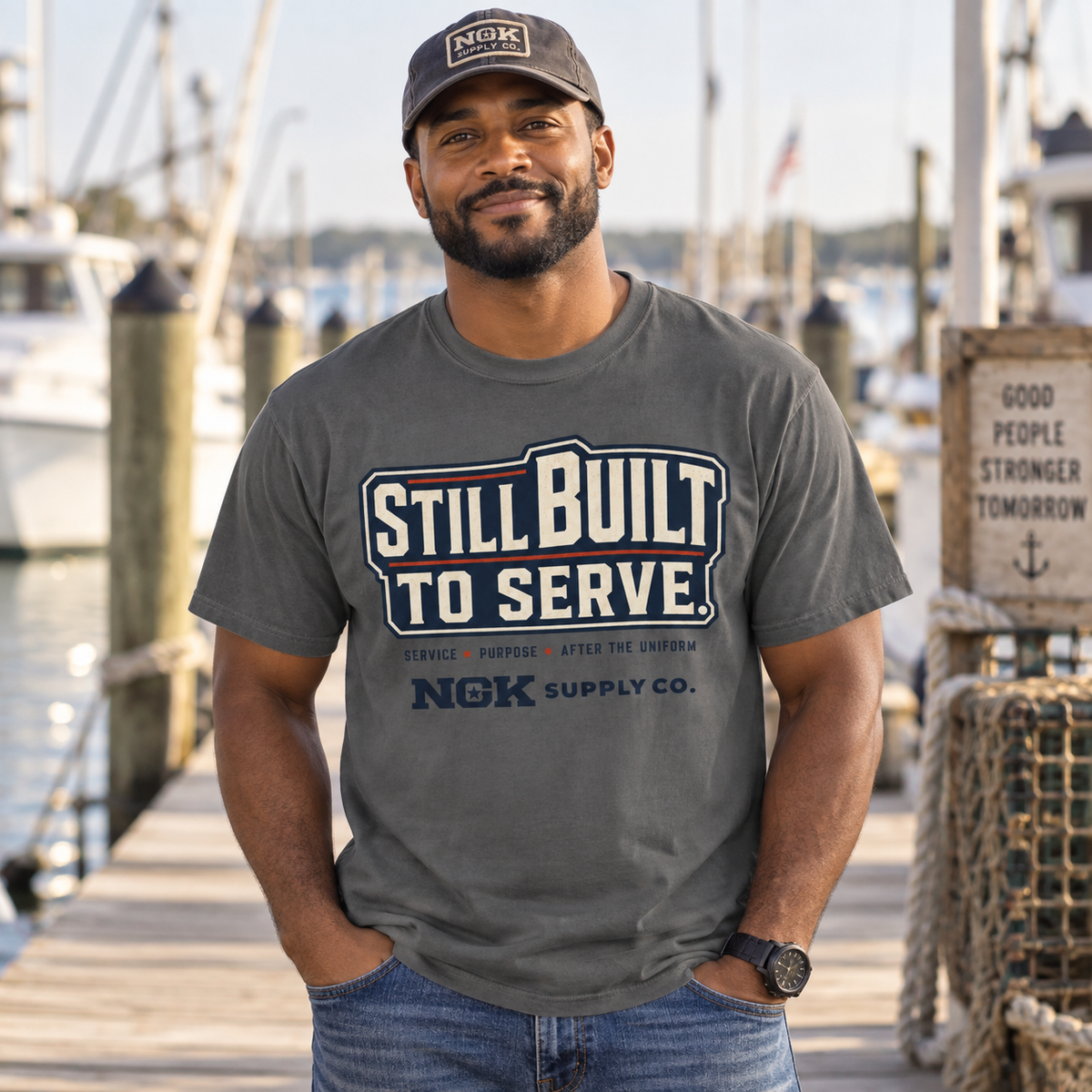 A man standing on a wooden dock wearing a dark gray t-shirt with a 'Still Built to Serve' graphic by NGK Supply Co. and a matching NGK cap, with boats and a marina in the background.