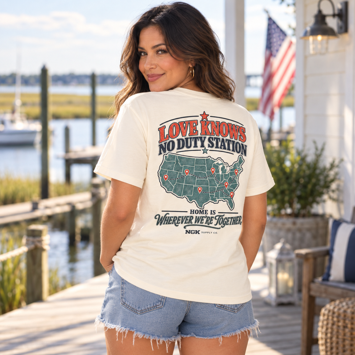 A woman stands on a wooden dock outdoors wearing a cream graphic T-shirt and denim shorts. The back of the shirt features a large map of the United States with red and navy text reading 'LOVE KNOWS NO DUTY STATION' and 'HOME IS WHEREVER WE'RE TOGETHER.