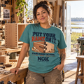 A woman stands in a woodworking shop wearing a teal graphic T-shirt with the text 'PUT YOUR NAME ON IT.' and an NGK woodworking illustration.